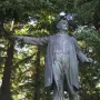 Harvey W. Scott statue of a man in a long coat pointing forward, set on a light-colored base among pine trees.