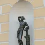 Apollino bronze nude male statue with one arm raised, standing in an arched niche beside a light yellow facade.