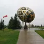 Vegreville Pysanka egg sculpture with geometric gold, black, and silver patterns in a park with trees and a pond nearby.