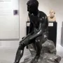 Seated Hermes statue of a nude male figure with winged sandals, shown on a rocky base in a museum display.