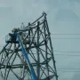 A large grey metal sculpture under construction with a worker in a blue boom lift nearby and power lines overhead.