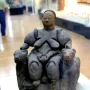 Seated Woman of Çatalhöyük figurine with animal heads on the armrests, displayed in a museum case.