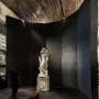 Michelangelo’s Rondanini Pietà displayed on a pedestal in a dim museum gallery, with a visitor standing nearby.