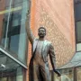 A bronze statue of A Lluís Millet in a formal suit and bow tie, with one arm raised and pointing upward.