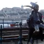 Little Princess bronze statue of a child sitting on a green railing with a city skyline and river in the background.