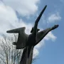 The Angel of Peace sculpture shown from below against a blue sky with clouds, with one arm extended and a pointed form rising beside it.