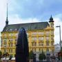The Brno astronomical clock stands in a sunlit city square with a woman in the foreground and market stalls and buildings behind it.