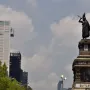 Cuauhtémoc Monument with a bronze figure in a feathered headdress on an ornate pedestal, set against modern buildings and trees.