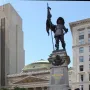 Maisonneuve Monument with a bronze figure holding a flag and sword atop a stone pedestal inscribed “MAISONNEUVE 1642,” with smaller figures below and city buildings in the background.