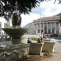 The Fountain of the Frogs stone fountain sprays water in a city square with a large classical building in the background.