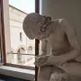 Boy with Thorn sculpture seen in profile, a light-colored boy leaning forward with his hands around his raised foot near a window.