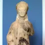 Chiotissa kore sculpture showing a female figure with braided hair and patterned draped garments on a red pedestal.