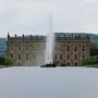 The Emperor Fountain shoots high in front of Chatsworth House, with green hills and trees under an overcast sky.