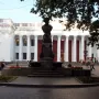Pushkin bust on a tall pedestal in a paved plaza before a grand columned building.