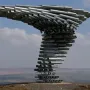 Singing Ringing Tree sculpture made of stacked silver metal pipes on a hillside with rolling landscape and distant wind turbines.
