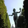 The Greek Actor statue on a stone pedestal with one arm raised holding a mask, set along a tree-lined path with a domed building in the distance.