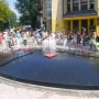 A small bronze Reksio monument beside a large fountain with water jets and people gathered around.