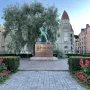 Aleksis Kivi Memorial bronze statue of a seated figure on a pedestal in a paved square, with hedges, red flowers, and buildings behind it.
