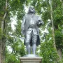 Benjamin Franklin statue in colonial attire on an ornate stone pedestal, framed by trees.