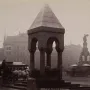 Michigan Soldiers\' and Sailors\' Monument beside an ornate stone canopy, with horse-drawn carriages and city buildings in the background.