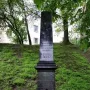 The Pit monument, a tall grey stone memorial with inscriptions and a carved emblem at the top, stands on a grassy hill with wreaths and flower baskets at its base.