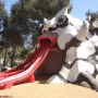 The Golem sculpture with a red mouth and three red slide chutes on sandy ground under a blue sky.