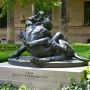 Dark bronze Hercules and the Nemean Lion statue on a stone pedestal beside a grand stone building.