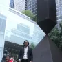 Broken Obelisk sculpture standing in front of modern glass buildings and greenery.