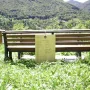 A wooden bench with a hanging plaque stands in a grassy area facing a green mountainside.