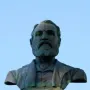 Weathered bronze bust of Maitland Brown in a suit and tie on a stone pedestal against a blue sky.