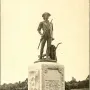 The Minute Man statue stands on a stone pedestal in a grassy area, holding a long gun with carved reliefs and inscriptions on the base.