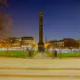 The Melville Monument rises above glowing lights in St Andrews Square at twilight, with illuminated buildings in the background.