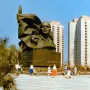 Ernst Thälmann Memorial in a wide public square with apartment buildings behind it and people walking nearby.