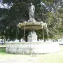 A classical draped female statue stands atop the Diana Fountain as water flows into a stone basin, with trees in the background.