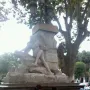 Ain El Fouara Fountain with a reclining nude female statue on a rocky base, framed by trees and a lamppost.