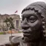Close-up of La Yunta bronze sculpture with a classical building and Peruvian flag in the background.