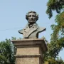 Dark bust of Pushkin on a stone pedestal outdoors, framed by trees against a blue sky.