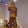 Rear view of the Sounion Kouros, a colossal ancient Greek stone statue of a nude male figure in a museum gallery.