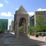 Bagley Memorial Fountain in a hazy city square, with a pyramidal stone top, arched openings, and ornate carvings.