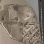 Fragmented relief from the Stele of the Vultures showing a large figure holding a staff with a bird emblem above smaller figures trapped in a net, with cuneiform text above.