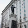 The Brigham Young Monument with a bronze figure atop a tall stone pedestal and relief sculptures at the base, with a white building and bare trees behind it.