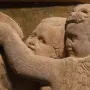 Close-up of a marble relief from the Pulpit of Prato Cathedral showing winged figures, faces, and a hand in front of a mosaic background.