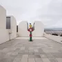 The Caress of a Bird sculpture with stacked green, red, and yellow shapes topped by a small blue bird in a courtyard.