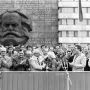 Karl Marx Monument beside a crowd gathered for an outdoor event, with a building facade behind it bearing a multilingual slogan.