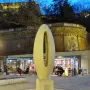 Zero Kilometre Stone on a paved plaza with a cafe and illuminated brick structure in the background.