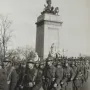 USS Maine National Monument with a sculptural figure and horses atop a tall stone base, seen behind marching soldiers.