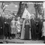 A dark Joan of Arc monument draped with garlands and foliage stands behind a cardinal and children in historical costumes.
