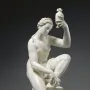 White sculpture of a kneeling female figure holding a small vessel aloft beside a draped pillar.