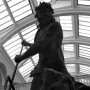 Neptune and Triton sculpture seen from below, with Neptune holding a trident and Triton blowing a conch shell beneath a glass ceiling.