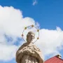 Statue of St. John of Nepomuk viewed from below against a blue sky, wearing a halo of stars and holding an object to his chest.